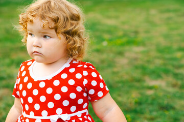 A young child with blonde curly hair gazes thoughtfully while wearing a bright red dress adorned with white polka dots The grassy background enhances the playful atmosphere of the moment