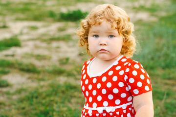 A young child with blonde curly hair gazes thoughtfully while wearing a bright red dress adorned with white polka dots The grassy background enhances the playful atmosphere of the moment