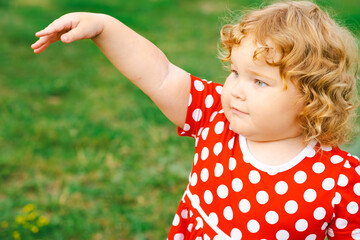 A young child with curly hair enjoys time outdoors wearing a bright polka dot dress while exploring the grassy area of a park in the afternoon sun