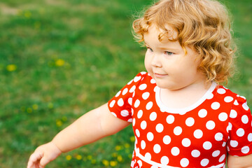 A young child with curly hair enjoys time outdoors wearing a bright polka dot dress while exploring the grassy area of a park in the afternoon sun