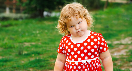 A child stands in a grassy area wearing a red dress with white polka dots showing an angry expression The surroundings are bright and sunny with green grass in the background