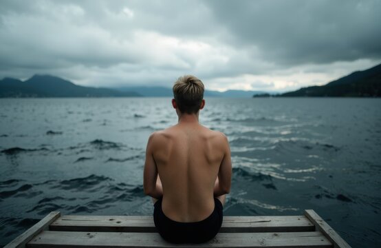 A young man sits shirtless on a dock overlooking a large body of water under a cloudy sky