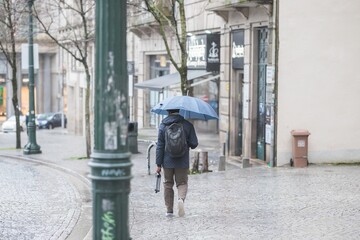 A man with a backpack walks away from the viewer, holding an umbrella against the rain. He is...