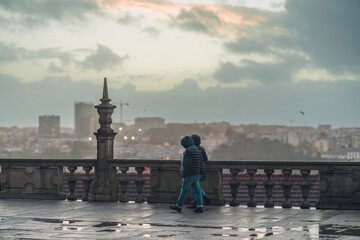 Two figures walk on the wet stone pavement of a balustrade in Porto. Rainwater is reflected on the ground. The cityscape is in the background under a cloudy sky