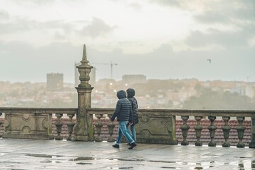 Obraz premium Two people with jackets walk along the stone balustrade overlooking Porto. The ground is wet, and the sky is overcast, creating a moody, pensive atmosphere in Vila Nova de Gaia