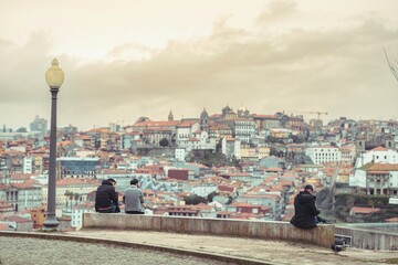 People sitting on a ledge admire the panoramic cityscape. Rooftops stretch into the distance beneath a cloudy sky in Vila Nova de Gaia, Porto. A street lamp stands tall nearby