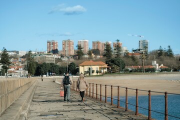 Two people walk away from the camera along a weathered pier in Porto. Buildings rise in the...