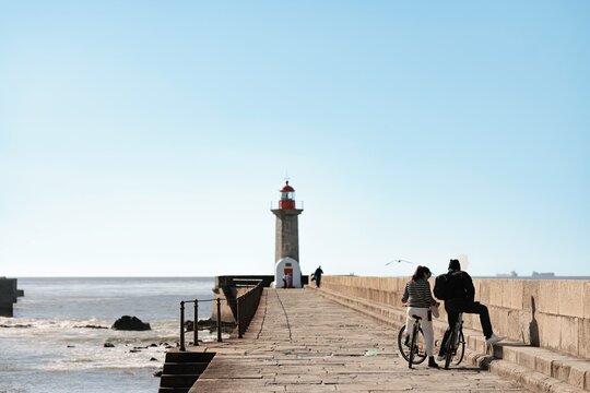 A couple stands with their bicycles on the pier leading to Farol de Felgueiras in Porto. Ocean waves crash against the pier, and a lighthouse stands at the end of the path