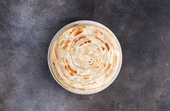 Traditional Indian paratha flatbread on white plate on a dark stone background. Top view.