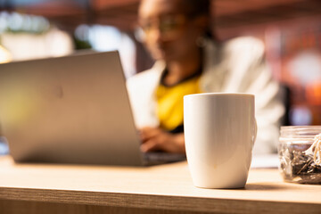 Focus on coffee mug on desk in front of remote worker typing on notebook keyboard. Close up of coffee cup on home office desk next to teleworker using laptop device to do job