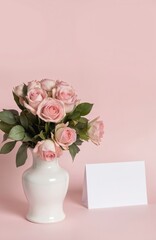 Obraz premium Delicate bouquet of pink roses in a white vase. A blank greeting card sits beside the floral arrangement on a soft pink background, perfect for Valentine Day or any romantic occasion.