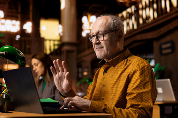 Elderly student attends a school webinar on laptop at a library, takes notes and does research during online lecture. Man studying remotely with a professor to obtain his diploma.