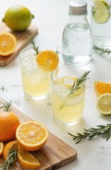 Fototapeta premium Refreshing homemade lemonade with fresh oranges, lemons, rosemary sprigs on white table. Two glasses filled with cold citrus drink, ice, fruit slices. Perfect summer beverage for healthy detox,