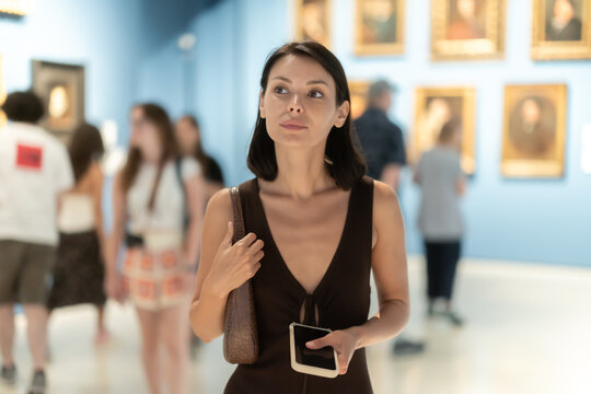 Female visitor with phone walks along painting exhibition pavilion. She inspects paintings, intellectual entertainment, European galleries attract tourists. Paintings in gold frames, Renaissance art - Powered by Adobe