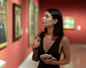 Young woman in a summer dress stands in art gallery with an audio guide in her hands. Modern ways...