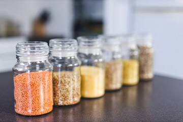 clear pantry jars with different types of grains and legumes including quinoa  lentils buckwheat and barley on benchtop with kitchen bokeh, simple ingredients