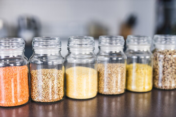 clear pantry jars with different types of grains and legumes including quinoa  lentils buckwheat and barley on benchtop with kitchen bokeh, simple ingredients