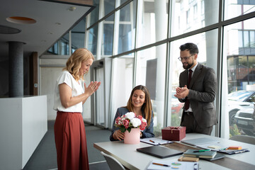 Business colleagues clapping while congratulating businesswoman receiving flowers and gift