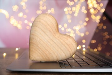 Online dating site. Wooden heart and laptop on table against blurred lights, closeup. Bokeh effect