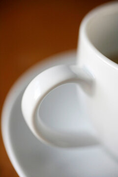 An artistic close-up of a clean, white coffee cup handle on a matching saucer, highlighting simple elegance and modern design with a warm, blurred background.