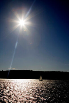 A serene sailboat glides across shimmering water under a brilliant sun with a striking lens flare. The peaceful silhouette contrasts beautifully with the bright sky.
