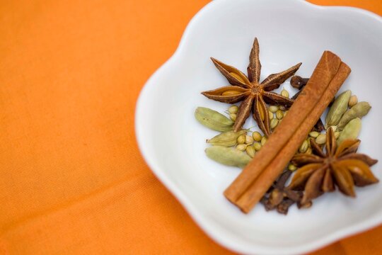 Close-up of whole star anise, cinnamon sticks, cardamom pods, and coriander seeds elegantly arranged in a white bowl on a bright orange fabric. ces