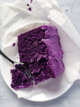 A rich slice of Ube cake with vibrant purple frosting, partially eaten with a fork on a white plate, presented on parchment paper over a marble background.