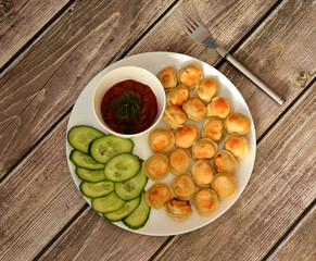 A plate of fried dumplings with fresh cucumber slices and tomato sauce on a wooden table, a fork nearby.