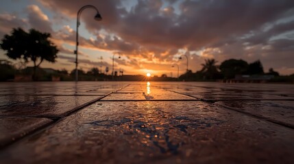 Dramatic sunset reflected on wet decorative pavement in an urban park, with silhouettes of trees and streetlights under a vibrant orange and purple sky filled with dynamic clouds.

