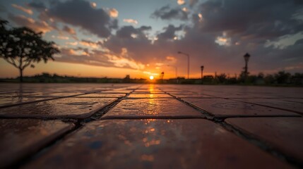 Dramatic sunset reflected on wet decorative pavement in an urban park, with silhouettes of trees and streetlights under a vibrant orange and purple sky filled with dynamic clouds.

