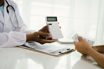 A doctor consults with a patient, explaining medical documents at a desk with a clipboard, laptop,...