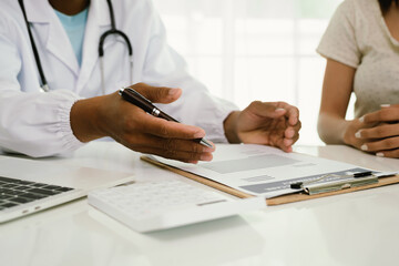 A doctor consults with a patient, explaining medical documents at a desk with a clipboard, laptop,...