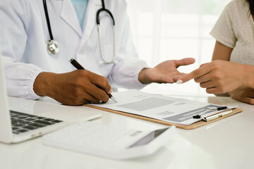 A doctor consults with a patient, explaining medical documents at a desk with a clipboard, laptop,...