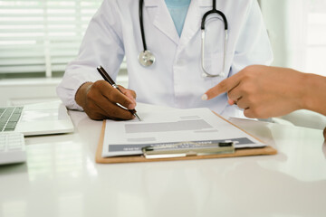 A doctor consults with a patient, explaining medical documents at a desk with a clipboard, laptop,...