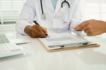 A doctor consults with a patient, explaining medical documents at a desk with a clipboard, laptop,...