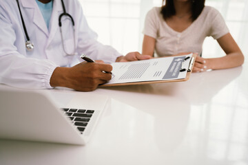 A doctor consults with a patient, explaining medical documents at a desk with a clipboard, laptop,...