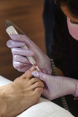 Close-up of a pedicurist wearing pink gloves removing gel polish from toenails using an electric...