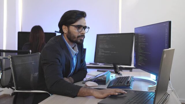 A focused Indian software engineer working on a laptop at his multi-monitor workstation in a modern tech office. Concept for deep work, programming, and data analysis.