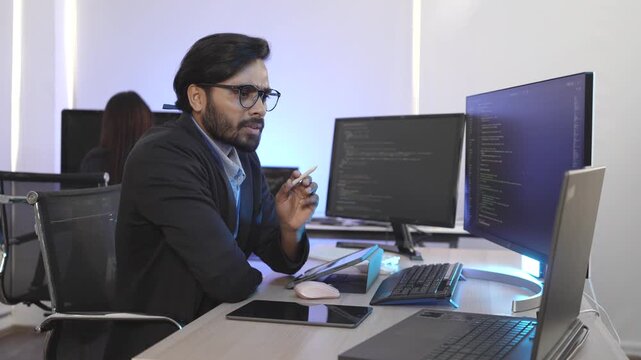 A focused Indian software engineer working on a laptop at his multi-monitor workstation in a modern tech office. Concept for deep work, programming, and data analysis.