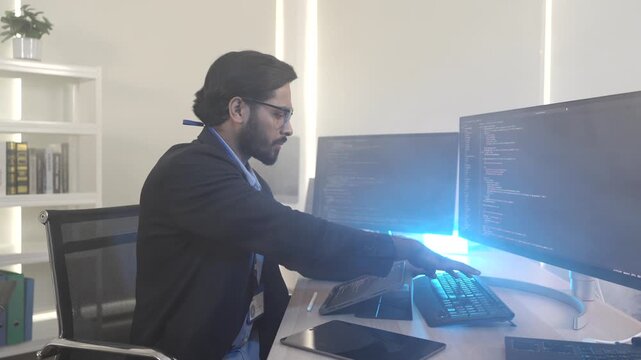 A focused Indian software engineer working on a laptop at his multi-monitor workstation in a modern tech office. Concept for deep work, programming, and data analysis.
