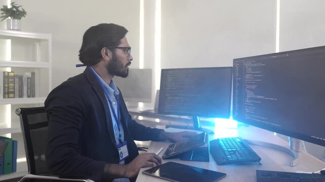 A focused Indian software engineer working on a laptop at his multi-monitor workstation in a modern tech office. Concept for deep work, programming, and data analysis.