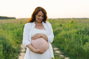 A pregnant woman stands in a green field. She gazes lovingly at her baby bump as the sun sets...