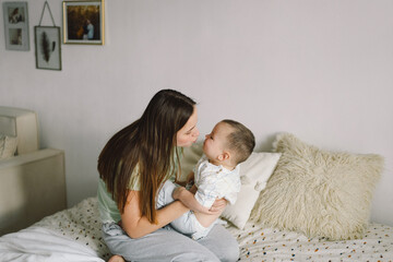 Home portrait of a baby boy with mother on the bed relaxing and playing in the bed at the weekend together, lazy morning, warm and cozy scene. Mom holding and kissing her child. Mother day concept