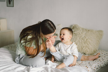 Home portrait of a baby boy with mother on the bed relaxing and playing in the bed at the weekend together, lazy morning, warm and cozy scene. Mom holding and kissing her child. Mother day concept