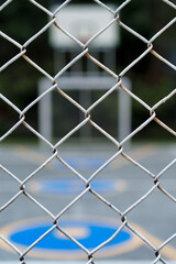 Street view of a multi-sport court with blue and yellow basketball and soccer markings, blurred in the background behind a white metal wire fence 