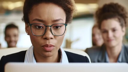 A focused professional woman wearing glasses concentrates on her laptop screen in a modern office environment.