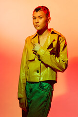 Young woman with trendy haircut wearing yellow jacket and green pants posing against vibrant orange background, showcasing modern fashion and bold colors.