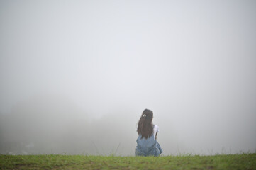 A solitary person with long hair sits on grass, facing a thick, gray fog that obscures the...