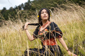 Indigenous woman sits in a field of tall grass, wearing a traditional embroidered dress and holding...
