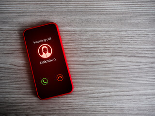 Malicious, abusive or threatening calls concept. Incoming call alert with unknown caller, scammer icon, unknown calling display showing on red smart mobile phone screen on wood table with copy space.